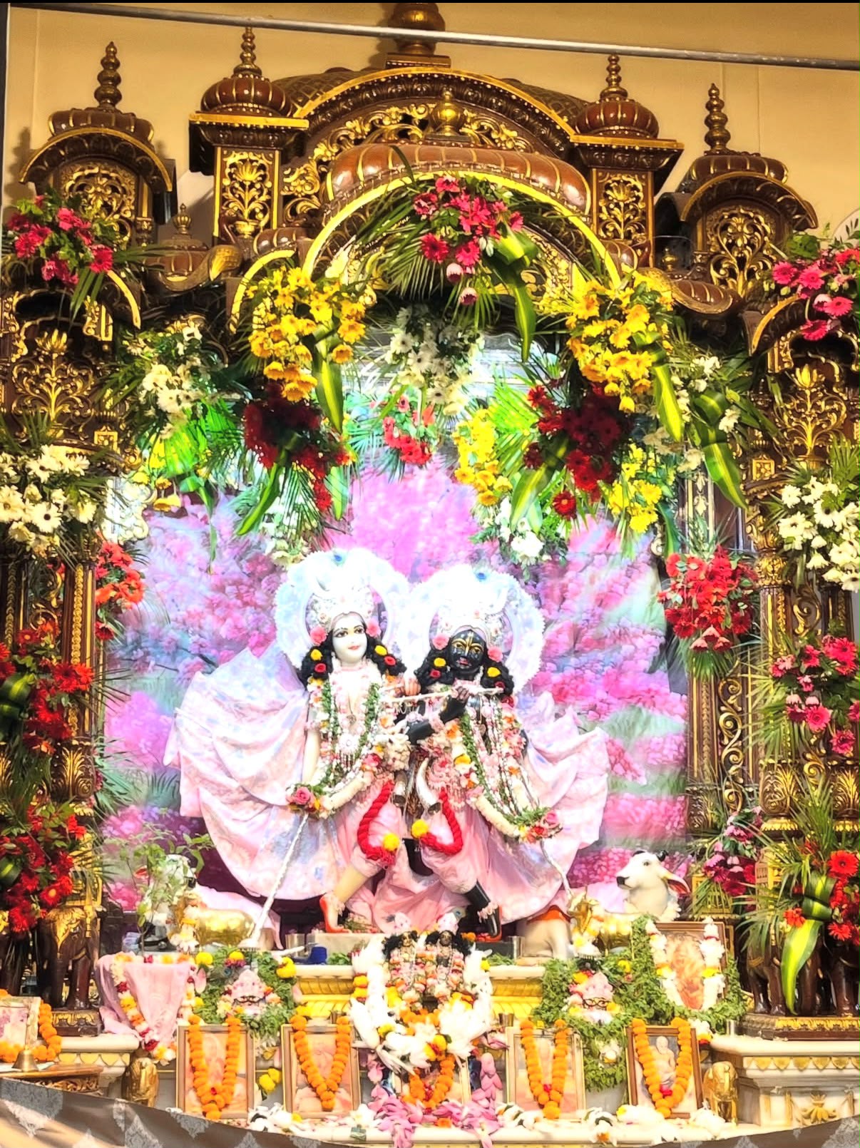 Radha and Krishna dressed in pink, surrounded by flowers at an ornate altar
